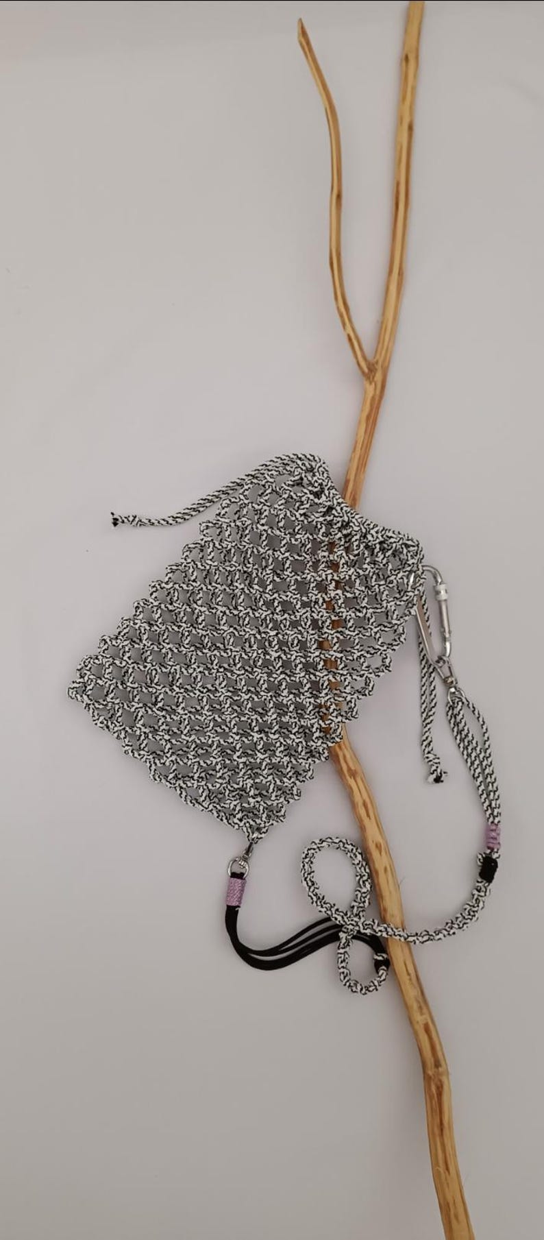 May include: A small, woven bag in black and white with a drawstring closure. The bag has a long strap with a silver clasp and a black and pink accent. A wooden branch is in the background.