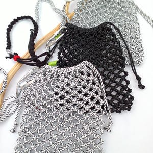 May include: Close-up of several macrame drawstring bags in black and white. The bags are made of a woven, open-weave material with a drawstring closure. The bags are attached to a silver carabiner clip. A black macrame bracelet is also visible.