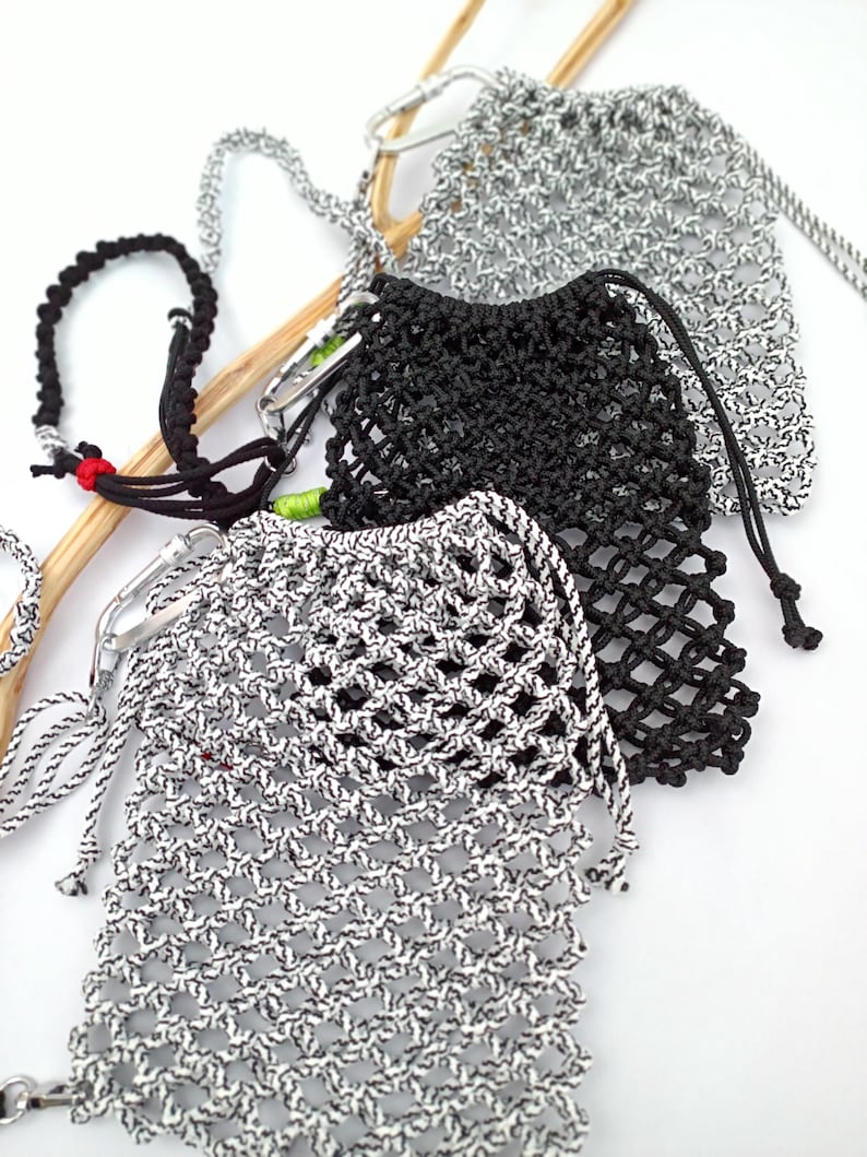 May include: Close-up of several macrame drawstring bags in black and white. The bags are made of a woven, open-weave material with a drawstring closure. The bags are attached to a silver carabiner clip. A black macrame bracelet is also visible.