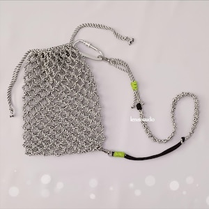 May include: A handmade, open-weave bag in black and white, with a drawstring closure. The bag has a matching rope strap with a carabiner clip and accents of neon green and black. The bag is designed for carrying small items.