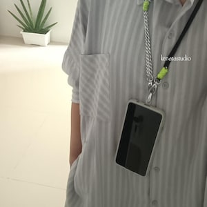 May include: A light gray, vertically striped shirt with a pocket and matching pants. A phone with a black screen is attached to a silver chain and black cord lanyard. The text "kenatastudio" is visible on the shirt.