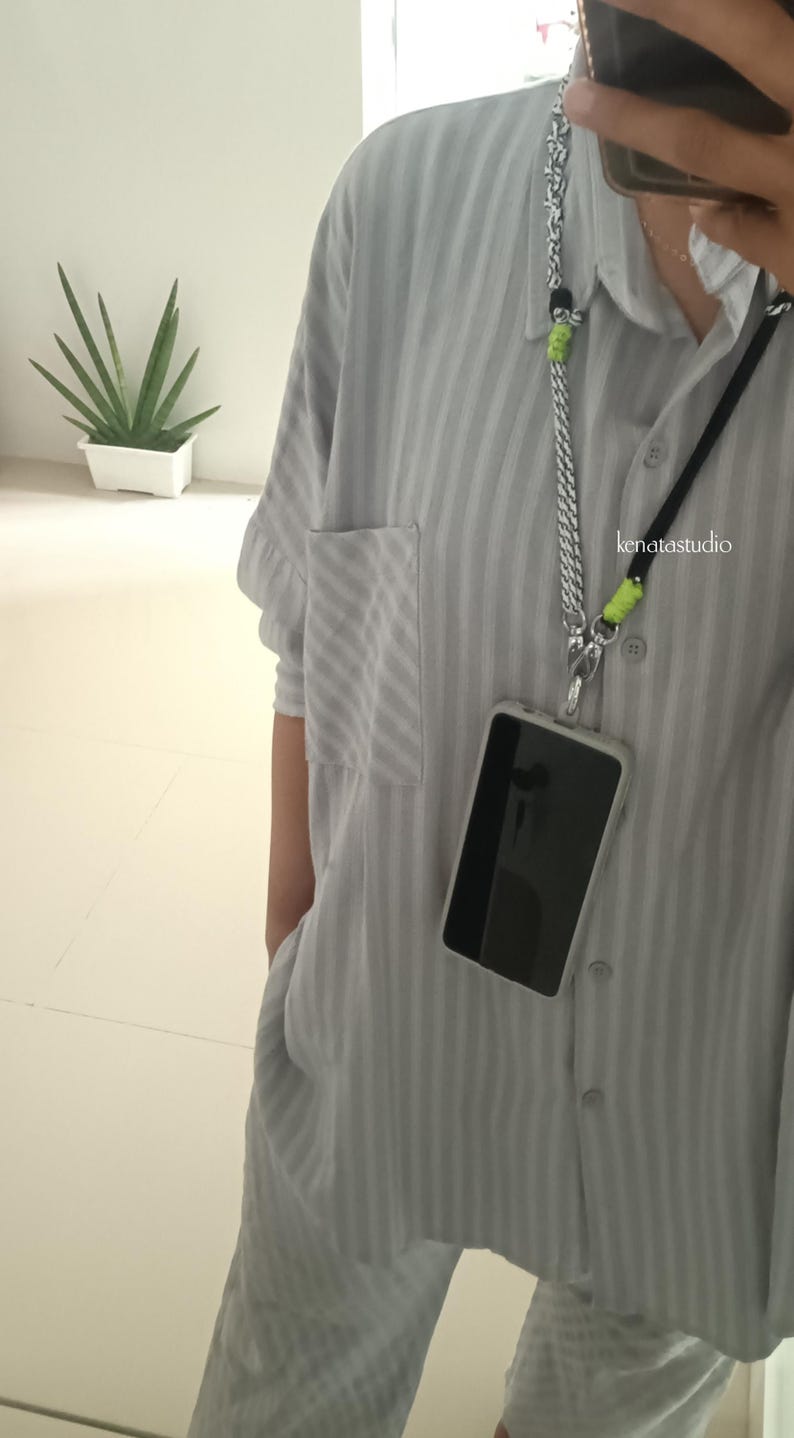 May include: A light gray, vertically striped shirt with a pocket and matching pants. A phone with a black screen is attached to a silver chain and black cord lanyard. The text "kenatastudio" is visible on the shirt.