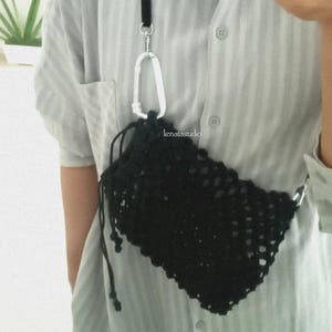 May include: Black crocheted crossbody bag with a drawstring closure, attached to a black strap with a silver carabiner clip. The strap has a red accent. The bag is worn over a light blue button-down shirt.