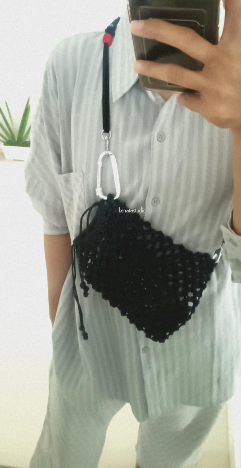 May include: Black crocheted crossbody bag with a drawstring closure, attached to a black strap with a silver carabiner clip. The strap has a red accent. The bag is worn over a light blue button-down shirt.