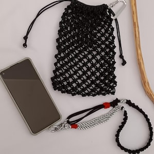 May include: Black macrame phone bag with a silver carabiner clip, a black lanyard, and a phone. The lanyard has a black and white braided section with red accents. The items are on a white surface.