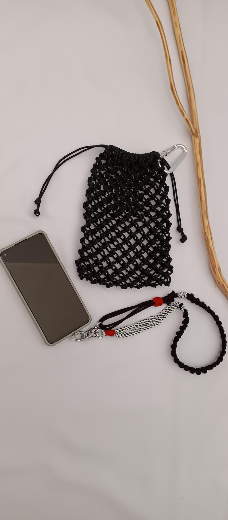 May include: Black macrame phone bag with a silver carabiner clip, a black lanyard, and a phone. The lanyard has a black and white braided section with red accents. The items are on a white surface.