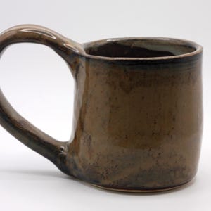 May include: A handmade brown ceramic mug with a large, curved handle. The mug has a glossy finish and a slightly irregular shape, suggesting it is handcrafted. The interior is a darker shade of brown. The mug is designed for hot beverages.