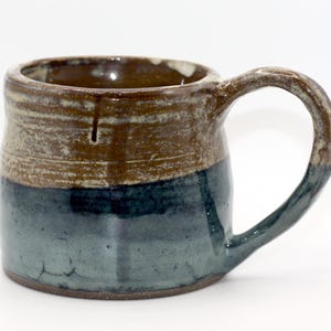 Puede incluir: Taza de cerámica hecha a mano con cuerpo redondeado y asa grande. La taza presenta un diseño de dos tonos, con una base azul oscuro y una sección superior marrón. El interior es de un marrón más oscuro. La taza es adecuada para bebidas frías o calientes.