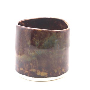 May include: A handmade ceramic cup with a unique, organic shape. The cup features a dark brown glaze with hints of green and blue, and a white base. The interior is dark, and the overall aesthetic is rustic and earthy.
