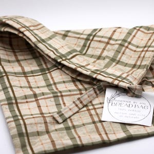 May include: A beige and green plaid bread bag made of 100% cotton. The bag is tied with a matching fabric tie and has a tag that reads "HANDMADE BY API BREAD BAG". The bag is designed to be machine washable on a delicate cycle.