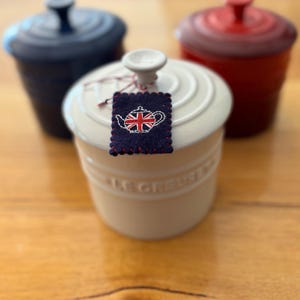 Puede incluir: Tres tarros de cerámica con tapa en color crema, azul oscuro y rojo. El tarro crema en primer plano tiene una pequeña etiqueta de fieltro rectangular azul oscuro con un diseño de tetera bordado en blanco que contiene la bandera británica.