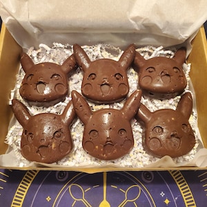May include: Six chocolate Pikachu-shaped brownies in a cardboard box lined with white paper shreds. The brownies are a rich brown color and have detailed facial features. The box sits on a purple and gold patterned surface.
