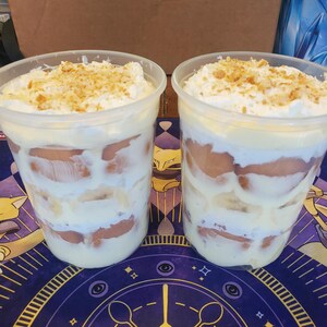 May include: Two clear plastic cups filled with layers of banana pudding, sliced bananas, and vanilla wafers, topped with whipped cream and crushed cookie crumbs. The dessert cups are positioned side by side on a dark blue patterned surface.