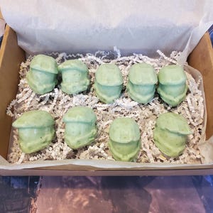 May include: A cardboard box containing nine light green, helmet-shaped molded treats nestled in white shredded paper filler. Each piece features a distinct visor and facial structure, arranged in two rows within the open container.