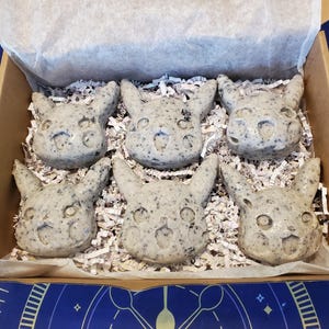May include: A cardboard box filled with six gray, speckled Pikachu-shaped soaps. The soaps are arranged on white crinkle paper. The box is lined with white paper and sits on a blue and purple patterned surface.