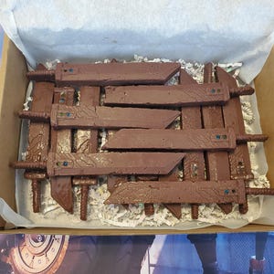 May include: A collection of brown chocolate sword-shaped treats arranged in a cardboard box. Each chocolate sword features a rectangular blade with a textured surface, a small handle, and a tiny embedded blue and green candy detail near the hilt.