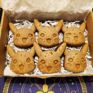May include: Six Pikachu-shaped cookies in a cardboard box with paper shred filler. The cookies are light brown with detailed facial features and ears. The box sits on a patterned surface with a blue and yellow design.