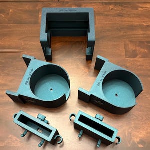 May include: An assortment of dark gray, 3D-printed plastic components, including a rectangular frame, two circular holders, and two smaller rectangular pieces. The parts are arranged on a wooden surface.