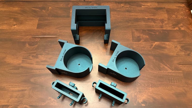 May include: An assortment of dark gray, 3D-printed plastic components, including a rectangular frame, two circular holders, and two smaller rectangular pieces. The parts are arranged on a wooden surface.