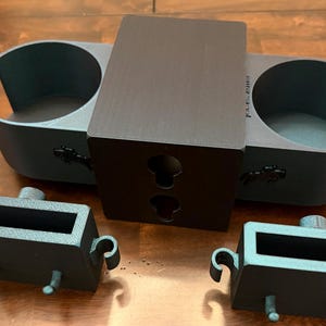 May include: A set of dark gray 3D-printed car accessories, including a dual cup holder, a central mounting piece with keyhole slots, and two rectangular storage compartments with handles and pegs. The items are displayed on a wooden surface.