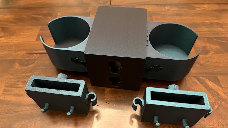 May include: A set of dark gray 3D-printed car accessories, including a dual cup holder, a central mounting piece with keyhole slots, and two rectangular storage compartments with handles and pegs. The items are displayed on a wooden surface.