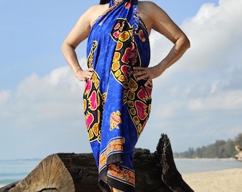 May include: A woman wearing a blue sarong with a colorful floral and turtle design. She is standing on a sandy beach with a large piece of driftwood behind her.