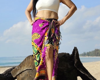 May include: A woman wearing a white crop top and a purple and black sarong with a floral and turtle design. She is standing on a sandy beach with a large piece of driftwood behind her.