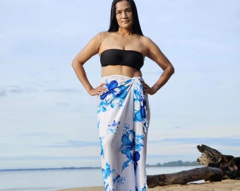 May include: A woman wearing a black tube top and a white sarong. The sarong features a blue floral pattern. The background shows a beach with sand, water, and a cloudy sky.