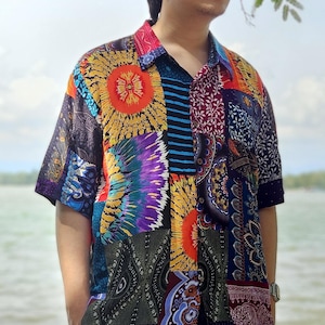 以下が含まれることがあります： カラフルなパッチワークのデザインの半袖ボタンダウンシャツを着た男性。シャツには、ペイズリー、花柄、幾何学模様など、さまざまなパターンが施されています。男性は黒いズボンを履いており、短い黒髪をしています。