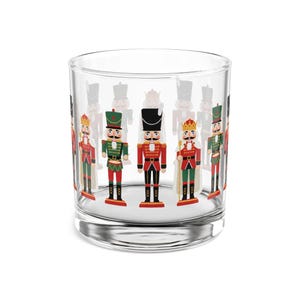 Op de afbeelding: Een helder glazen tumbler versierd met een herhalend patroon van kleurrijke speelgoed soldaat notenkrakers. De notenkrakers zijn afgebeeld in verschillende uniformen met rode, groene en zwarte jassen en gouden kronen. Het glas is cilindrisch met een dikke basis.