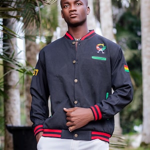 May include: Black bomber jacket with red and white striped cuffs and collar. Features a colorful emblem and the number 57 in yellow. The jacket has a green accent and a small flag on the sleeve.