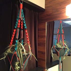 May include: A macrame plant hanger with a teal and orange design hangs from the ceiling with a potted plant inside. The hanger is made of knotted string with black beads.