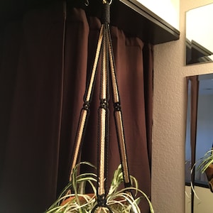 May include: A macrame plant hanger with a beige and black rope design, hanging from a ceiling hook. A green plant is in a brown pot inside the hanger.