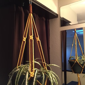 May include: A brown and yellow macrame plant hanger with a green plant in a black pot.