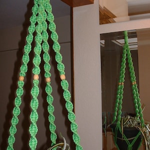 May include: Green macrame plant hanger with wooden beads. The hanger is made of twisted rope and has a classic design. It is hanging from a wooden beam.