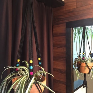 May include: A macrame plant hanger with colorful beads hangs from a hook in a room with brown wood paneling. A green plant with white stripes is in the pot.