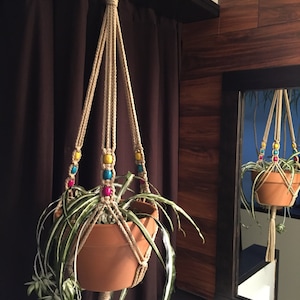 May include: A macrame plant hanger with colorful beads hangs from the ceiling, holding a terracotta pot with a green plant. The hanger is made of natural fiber rope and has a simple, bohemian style.