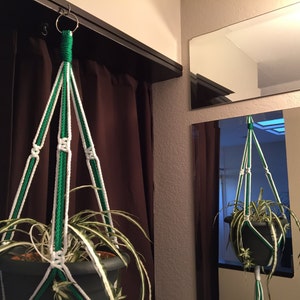 May include: A green and white macrame plant hanger with a potted plant hanging from it.