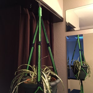 May include: A green and black macrame plant hanger with a potted plant hanging from it. The hanger is made of thick rope and has a knot at the top for hanging.