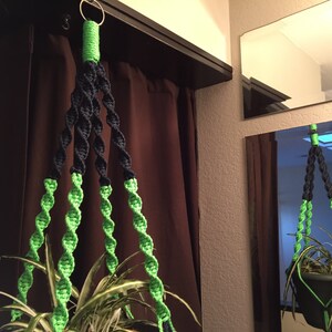 May include: A green and black macrame plant hanger with a metal ring at the top. The hanger is hanging from a ceiling and has a plant in a pot at the bottom.