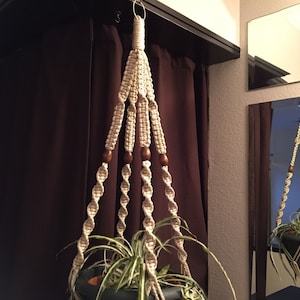 May include: A macrame plant hanger with wooden beads, hanging from a ceiling hook. The hanger is made of natural colored cord and has a spiral design. A green plant is hanging in the pot.