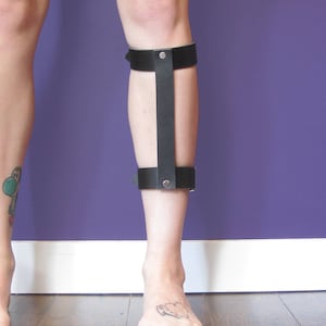 Puede incluir: Una férula de pierna de cuero negro con una sola correa en la parte delantera. La férula está asegurada con remaches metálicos.