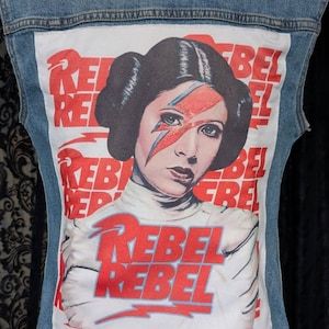 May include: A blue denim vest featuring a large rectangular white patch on the back. The patch displays a graphic of a person with dark hair in buns, a red lightning bolt across the face, and the repeated red text REBEL REBEL.