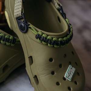 Puede incluir: Zuecos verde oliva con detalles en negro y verde neón. Los zuecos tienen una correa con un diseño tejido y un dije que dice "Money Maker". Los zapatos tienen múltiples orificios de ventilación.