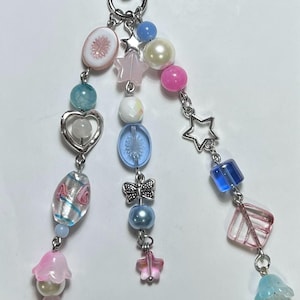May include: A beaded keychain with a silver clasp. The keychain features various beads in pastel colors, including pink, blue, and white, with star, heart, and butterfly charms. The beads are strung on silver chains.