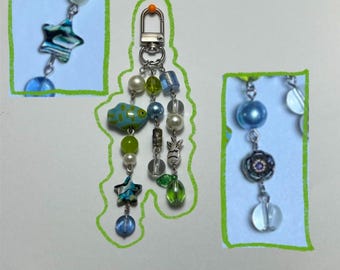 May include: A handmade keychain with a silver clasp and a variety of colorful beads. The beads include pearls, blue and green glass beads, a blue fish, a star, and a flower charm. The keychain is a unique and decorative accessory.
