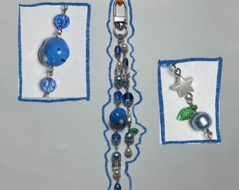 May include: A collection of handmade keychains and charms. The keychains feature blue beads, pearls, and charms, including a blue whale, a star, and a blueberry. The designs are set against a white background with blue outlines.