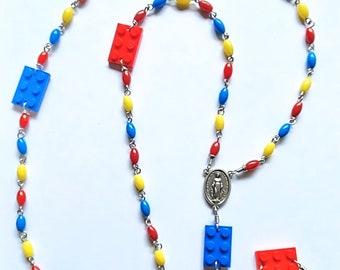 May include: A rosary featuring a silver-toned crucifix and a central medal depicting a figure. The chain consists of alternating oval beads in red, yellow, and blue, interspersed with rectangular plastic building blocks in the same primary colors.