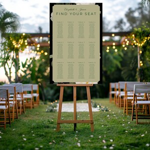 May include: A wooden easel displays a seating chart for Elizabeth & James' wedding. The chart has a black border and a light green interior with guest names organized by table number. The background shows an outdoor wedding setting with chairs and greenery.