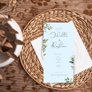 May include: A light blue wedding program with the names "Juliette & Alastair" in elegant script. The program features floral accents and the words "Welcome to the Wedding of" and "Thank You". It rests on a woven placemat.
